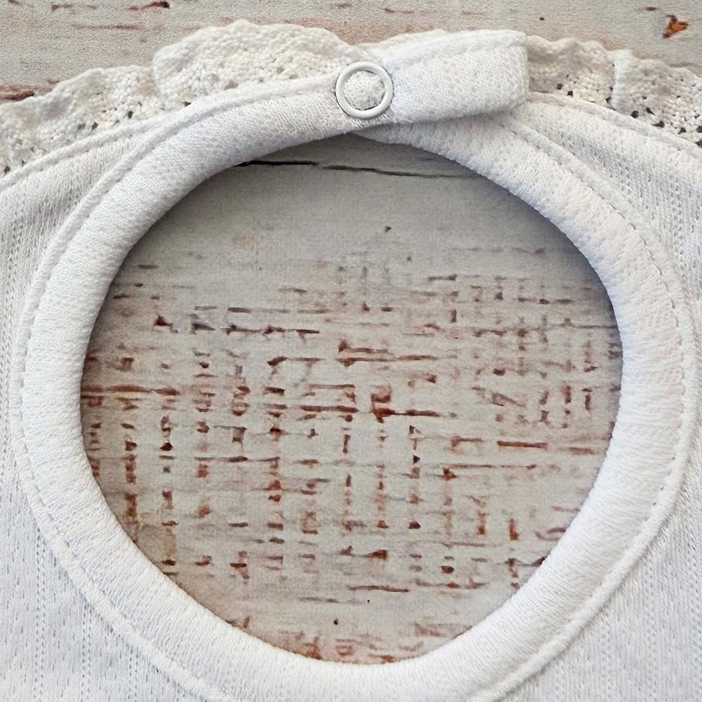Babero de niña blanco con ojal circular y volante calado en el contorno del cuello.